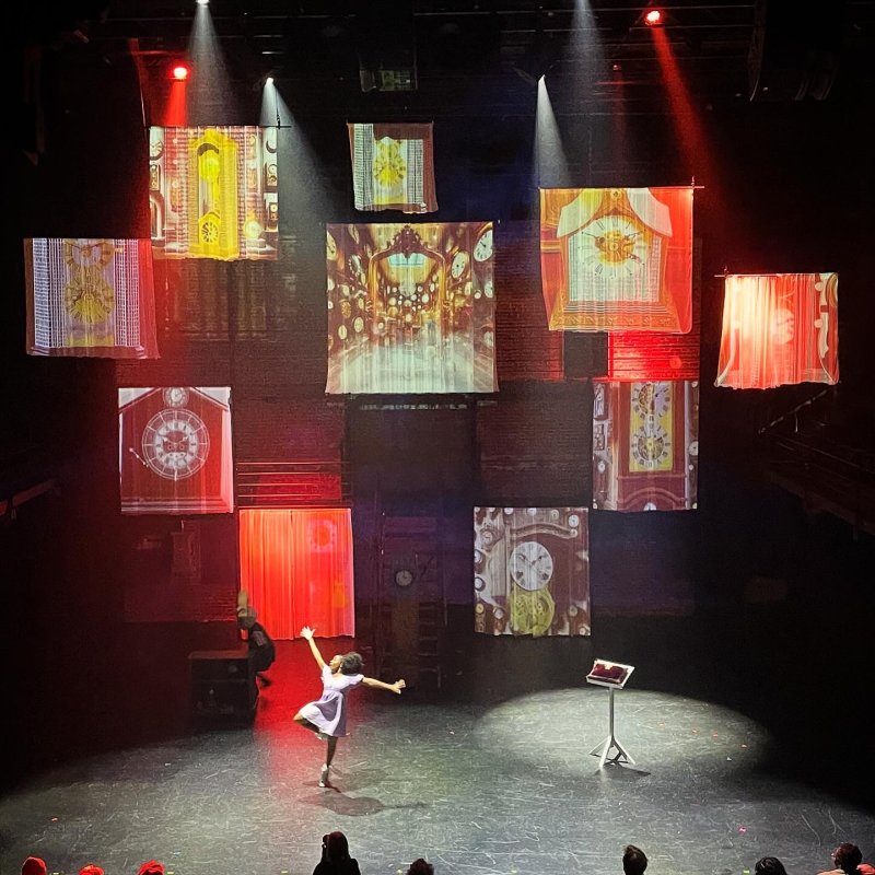 Stage performance with dancer, clock projections on fabric, red and white spotlights.
