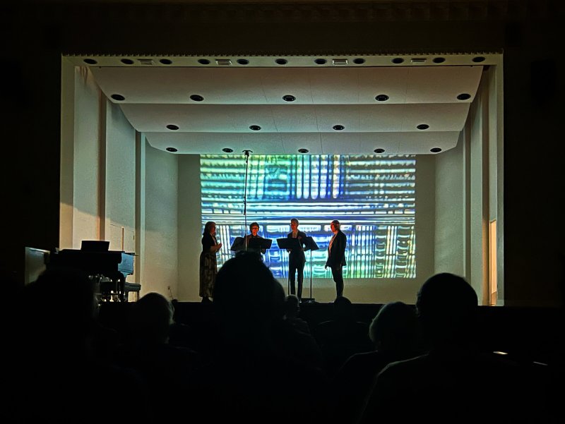 Four musicians on stage with a large screen behind them displaying abstract blue and green patterns.