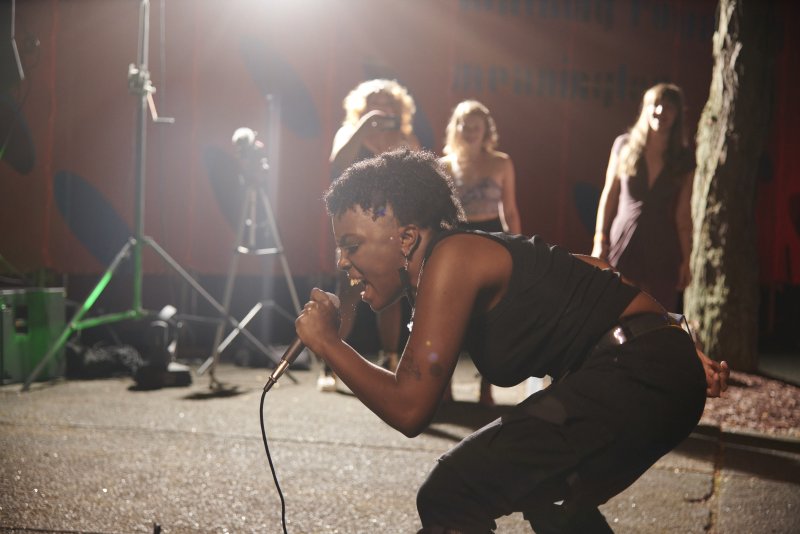 Outdoor performance scene with a Black singer in a black outfit singing into a microphone, leaning forward with mouth open. Soft focus background with other people and stage equipment. Bright stage lighting.
