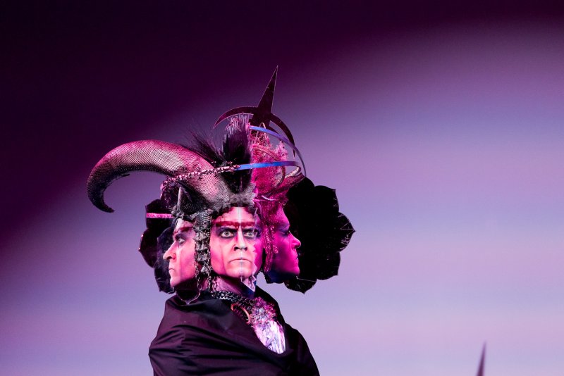 Performer Darrell Thorne in elaborate costume, featuring multiple faces, horns, and spikes, against a purple and pink gradient background.