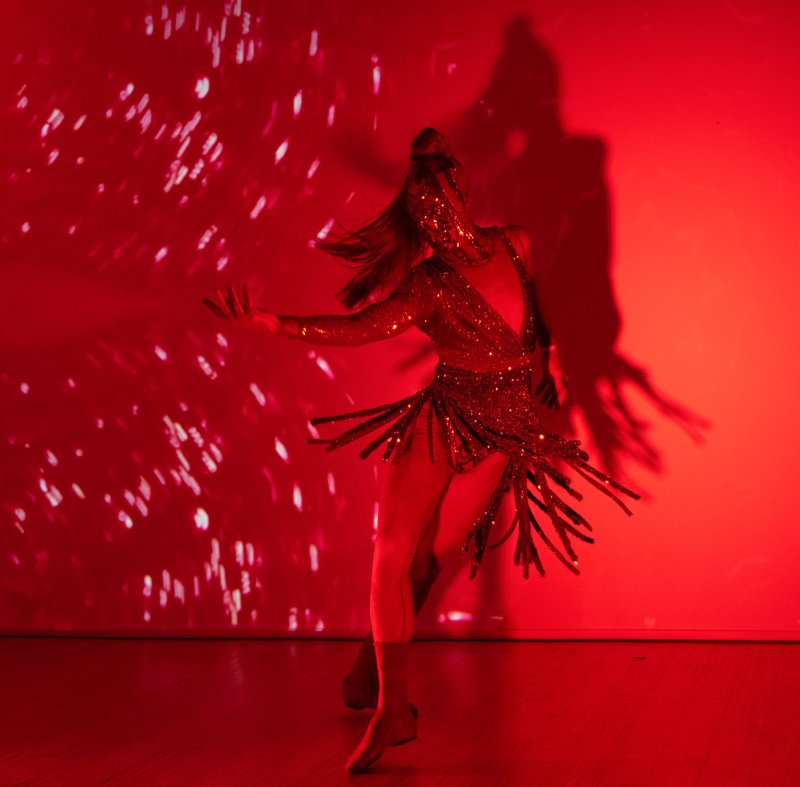 Dancer in a sparkling red costume with fringe, illuminated by red light and a projected pattern of white specks, casting a large shadow.