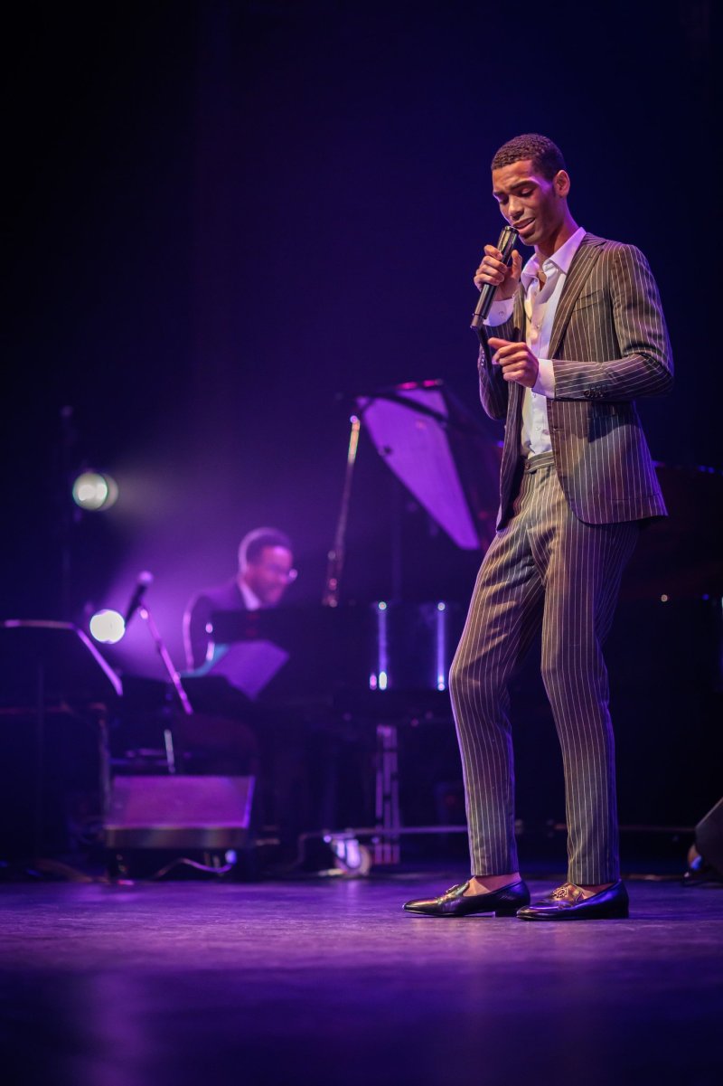 A male singer in a pinstripe suit and loafers performs on stage, holding a microphone under purple lights, with a pianist in the background.