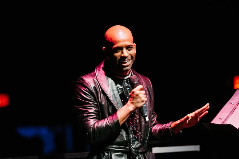 Bald person in a black leather jacket and sequined shirt, holding a microphone, smiling on a dark stage with pink light.