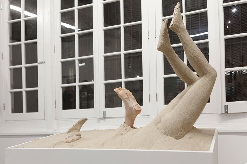Sculpture of human legs and feet emerging from sand in a white box, set against large white-framed windows.