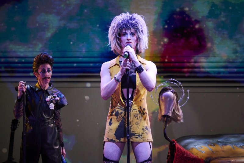 Drag queen in yellow tunic and fishnets sings into a microphone, another person in leather and fishnets stands beside them.