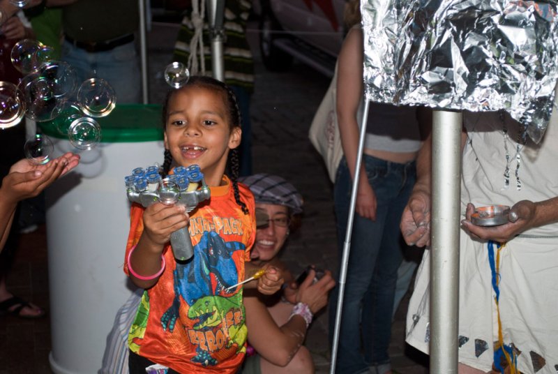 A young girl with braids smiles while holding a bubble gun, surrounded by bubbles and people.