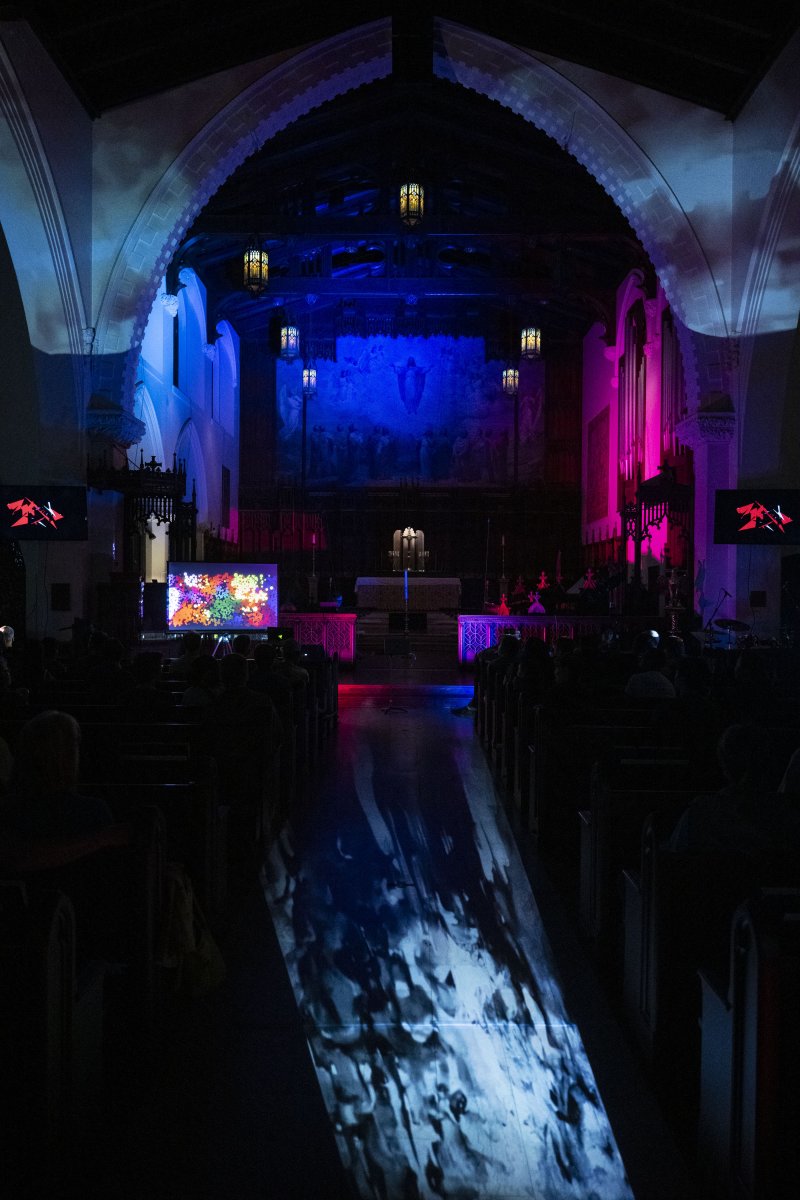 Dark church interior with blue and pink lights illuminating walls, a colorful screen on the left, and a projected pattern on the aisle.