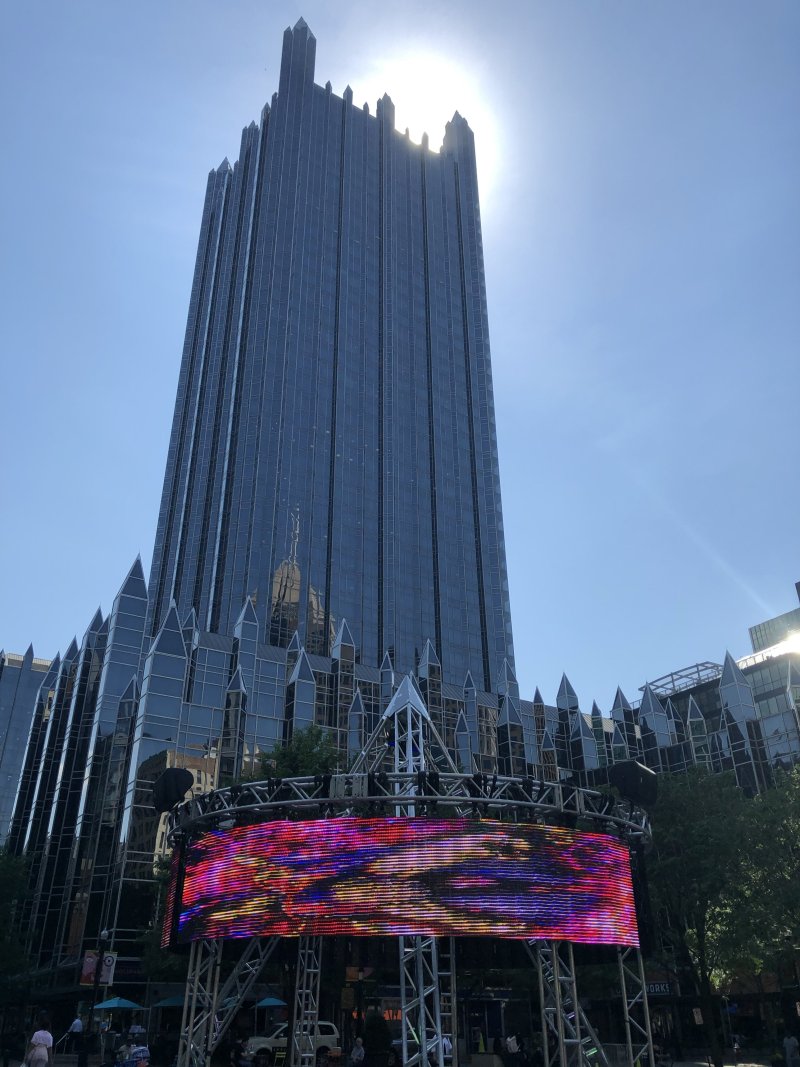 Tall, dark glass skyscraper with pointed spires rises above a large, horizontal LED screen displaying vibrant pink, red, and blue abstract patterns. Blue sky and sunlight in background.