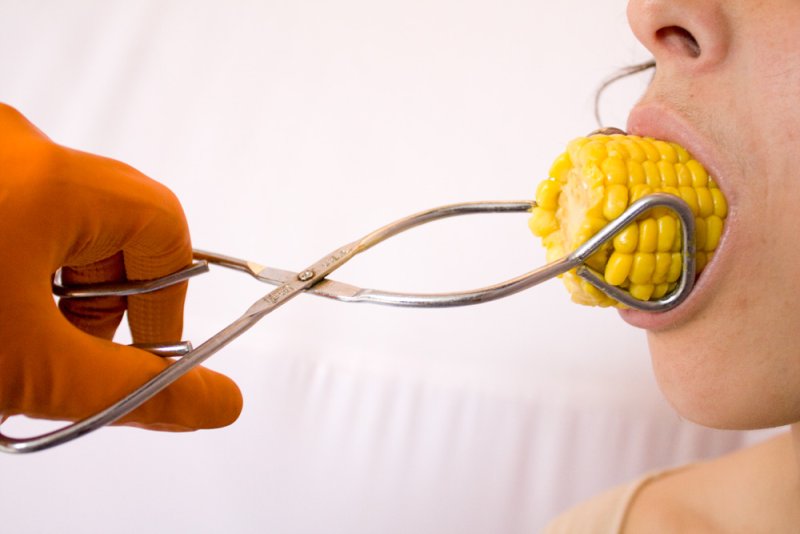 A person's mouth holds a corn cob, while an orange-gloved hand uses metal tongs to hold the corn.