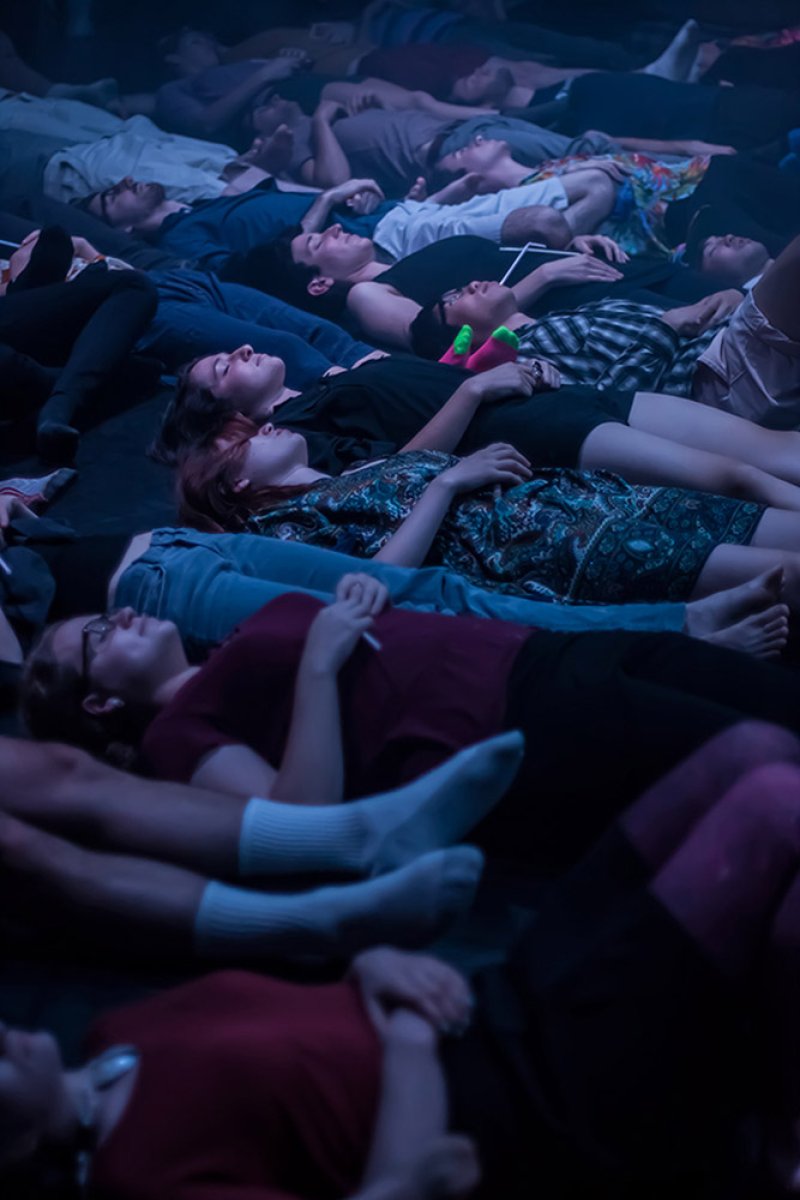 A group of people lie on their backs on a dark floor, illuminated by dim, cool light.