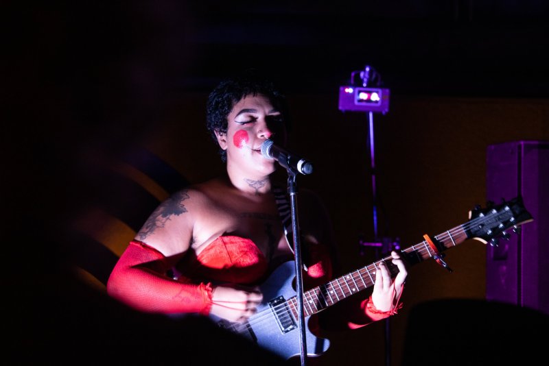 Darkly lit performance shot of a musician with red face paint singing into a microphone while playing a blue electric guitar. They wear red fishnet sleeves and a matching top. Stage lights illuminate the scene.