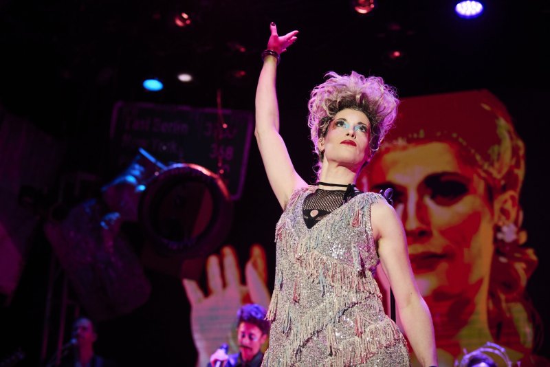 Drag performer in a sequined dress and blonde wig, arm raised, on a dark stage with red and blue lights and a projected image.