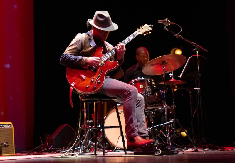 A man in a fedora plays a red electric guitar while seated on a stool, with a drummer in the background.