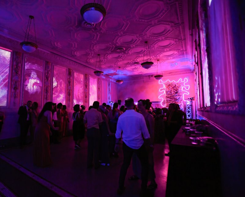 People gather in a large room bathed in purple and pink light, with ornate ceilings and abstract projections on the walls.