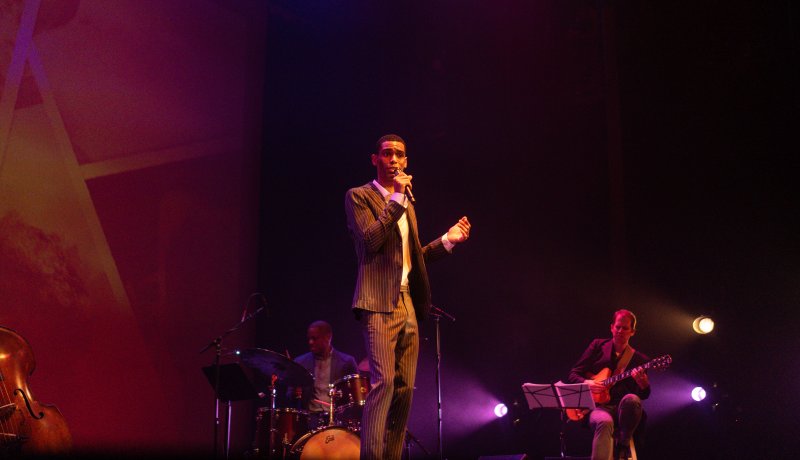 A singer in a striped suit performs on stage with a drummer and guitarist under purple and red lights.