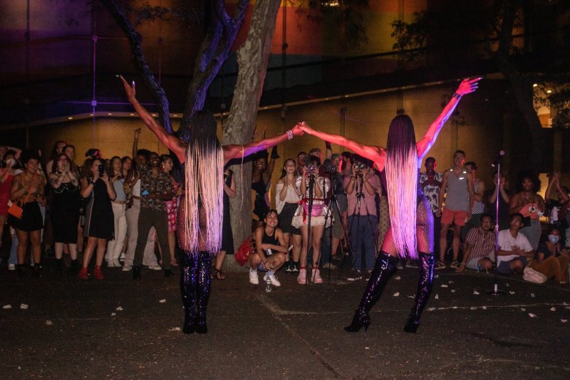Two performers in long blonde wigs and shiny black boots stand with arms raised before a crowd. Purple and red lighting illuminates the scene.