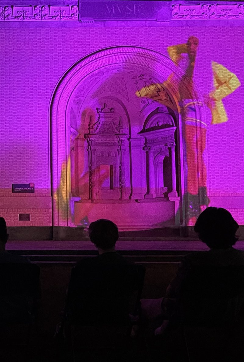 Purple and yellow light projection on a building facade with an arched entrance, viewed from behind three silhouetted figures.