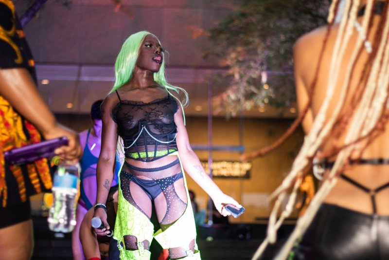Performance shot featuring a Black person with bright green hair wearing black lingerie and neon green chaps, holding a microphone. Other performers are partially visible.