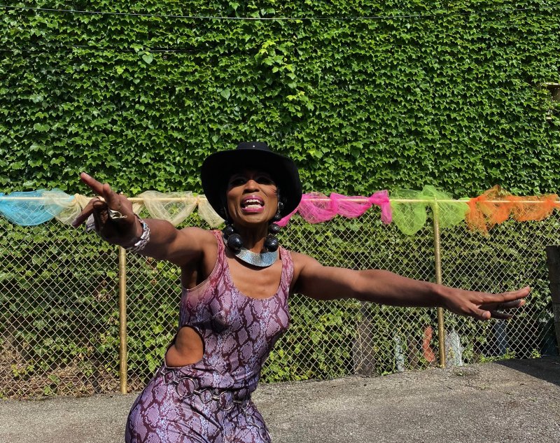 Drag queen in a purple snakeskin outfit, black hat, and silver jewelry, arms outstretched, against an ivy wall and chain-link fence.
