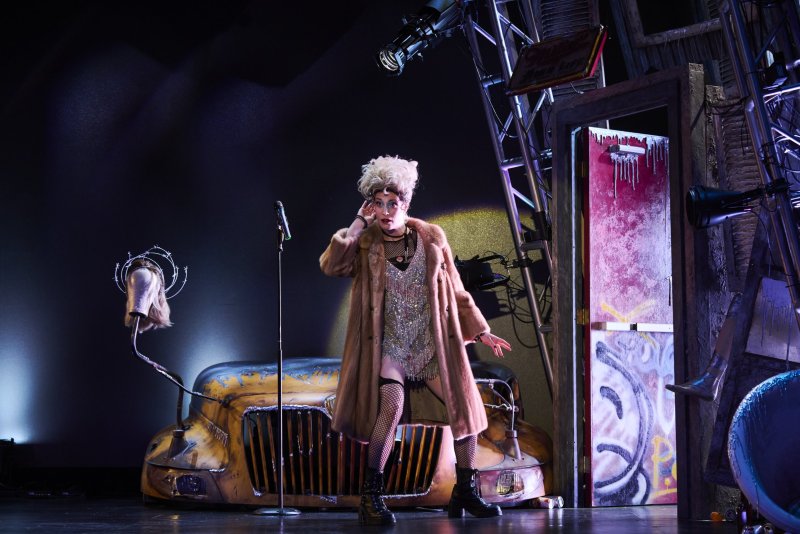 Drag queen in blonde wig, fur coat, and fishnets sings into a mic on a dark stage with a rusty yellow truck and a graffiti-covered door.