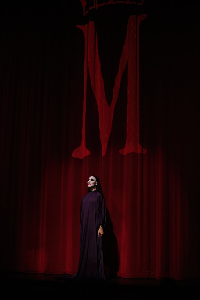 Figure in purple gown stands before red curtain with large red "M" above.