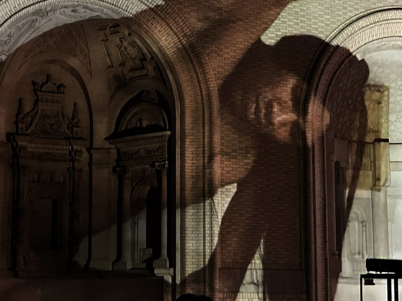 Shadow of a winged figure projected onto a brick archway and ornate stone wall, with warm brown and orange tones.