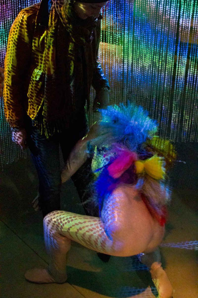 Performer in iridescent scale-patterned suit with blue, pink, and yellow feather boas kneels before a standing figure.