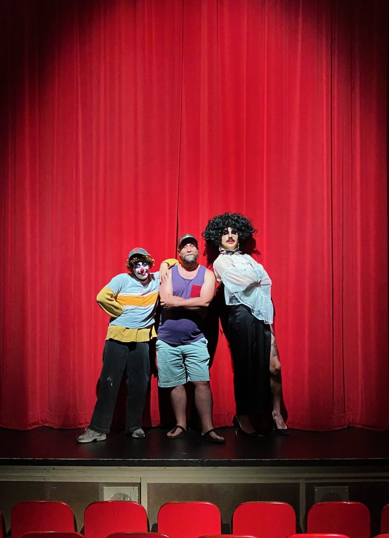 Three performers, one in clown makeup, one in drag, and one in casual wear, stand arm-in-arm on a stage before a red curtain.