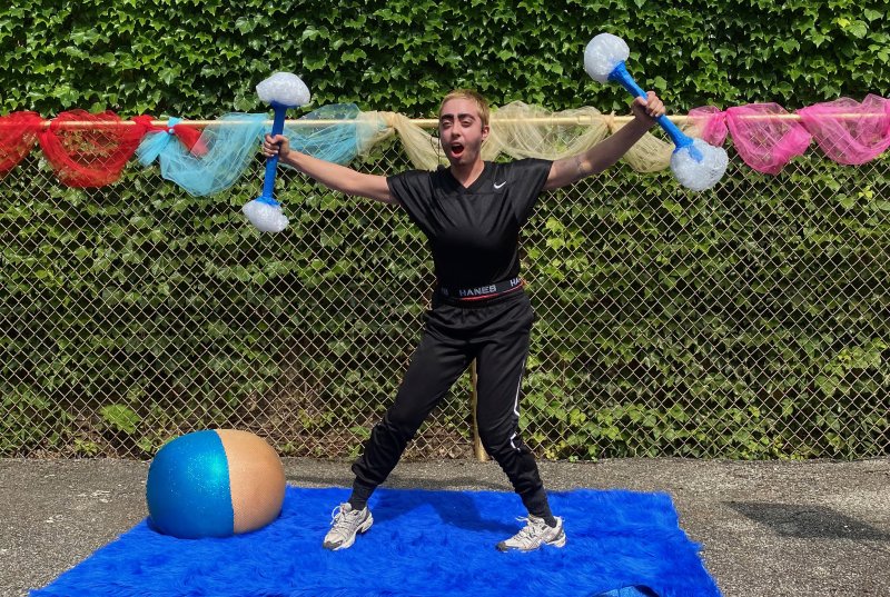 Person in black athletic wear holding blue and white dumbbells, standing on a blue shag rug in front of a fence with colorful fabric.