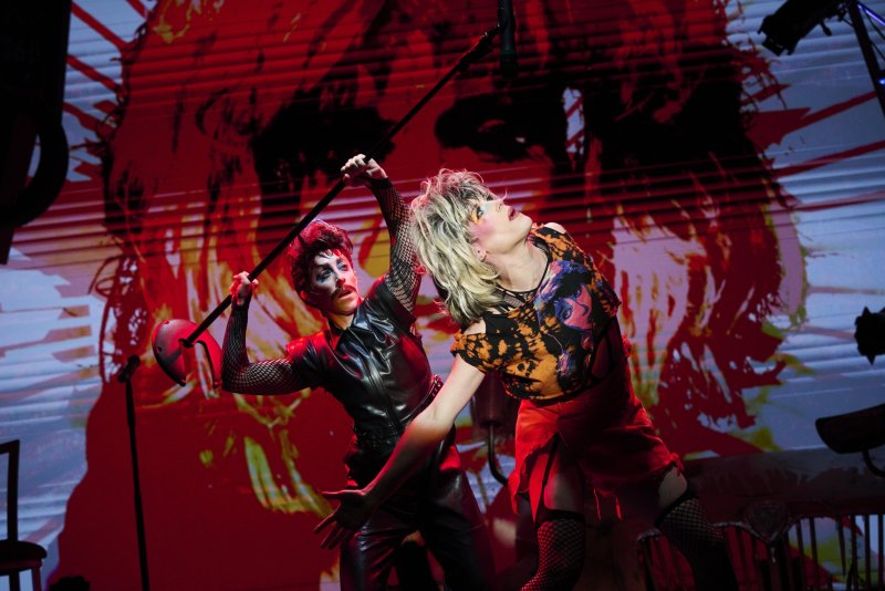 Two performers on stage, one in black leather and fishnets holding a mic stand, the other in a ripped top and skirt, both with dramatic makeup, against a red and black graphic back