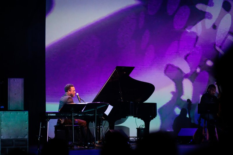 Scott N Andrew's "Suite Life" (2022-2023): A man plays a grand piano on stage, illuminated by a purple-hued projection.