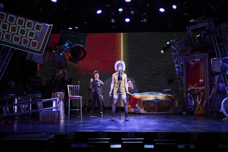 Two performers on a stage with a neon sign, a wrecked car, and a large screen displaying a red and green gradient.