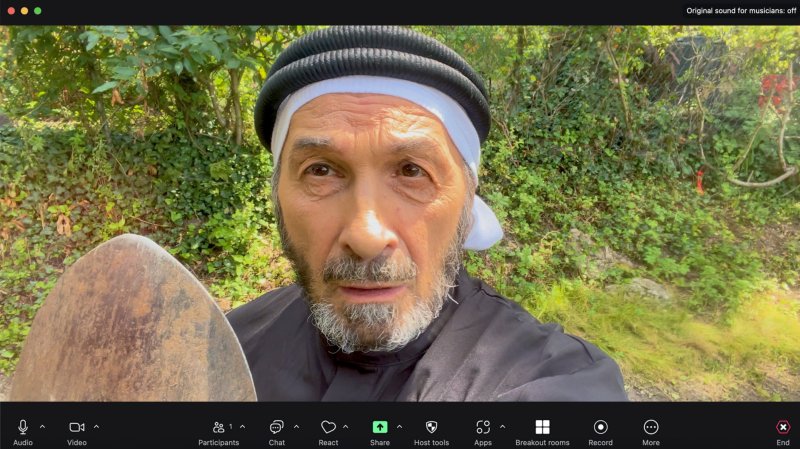 Man in black and white head covering, holding a shovel, looking at camera in a video call interface with green foliage background.