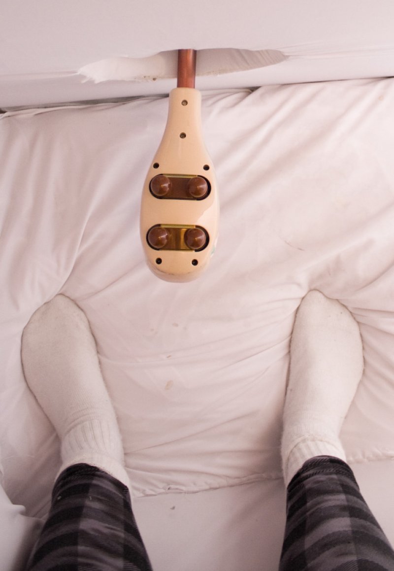Feet in white socks and plaid pants lie on a white sheet below a beige massager hanging from a copper pipe.