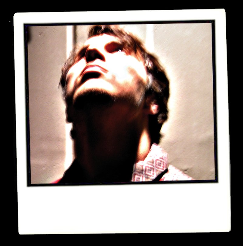 Polaroid of a man looking up, with dark shadows on his face and neck, wearing a patterned shirt.