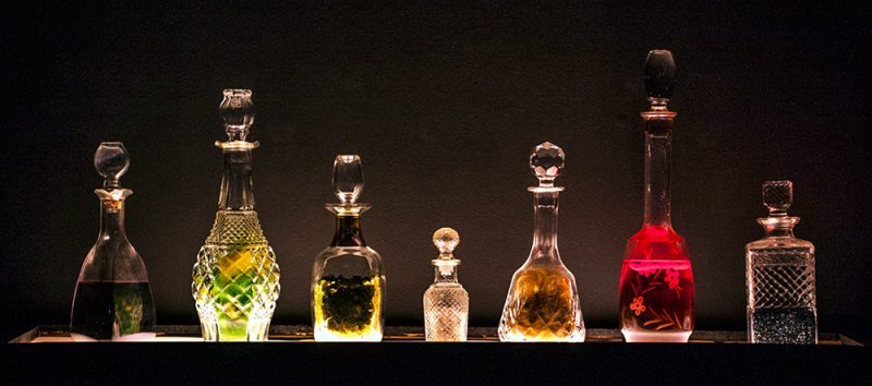 Seven ornate glass decanters, varying in size and shape, filled with colorful liquids, illuminated against a dark background.