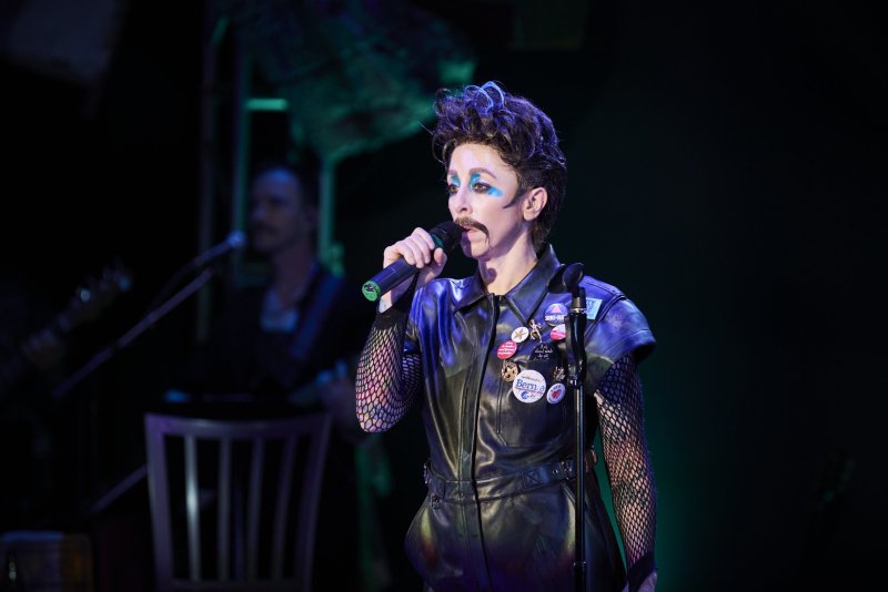 Performer with dark spiky hair, painted mustache, and blue eyeshadow, wearing a black leather vest over fishnet sleeves, singing into a microphone.