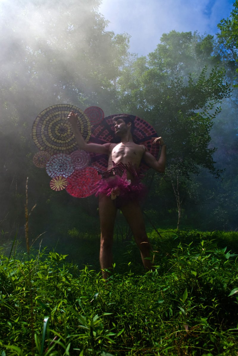 A shirtless person in a pink tutu and large, colorful circular wings stands in a misty forest.