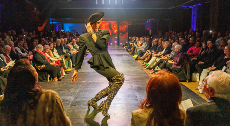Drag queen in black peplum top, pants, beret, and patterned boots striking a pose on a runway, with an audience seated on both sides.
