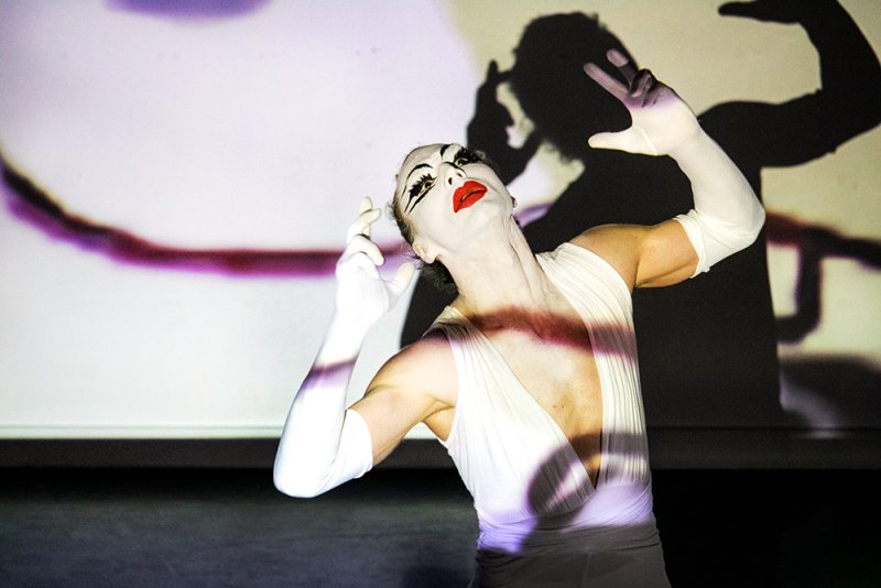 Performer in white mime makeup and gloves, red lips, white dress, with projected shadow on a light wall.