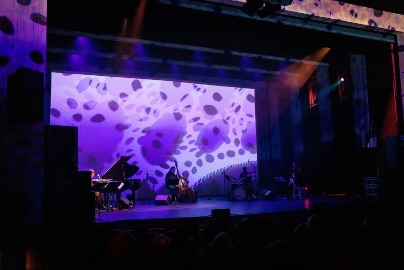 A band performs on a stage with a large screen displaying abstract purple and white shapes, under purple lighting.