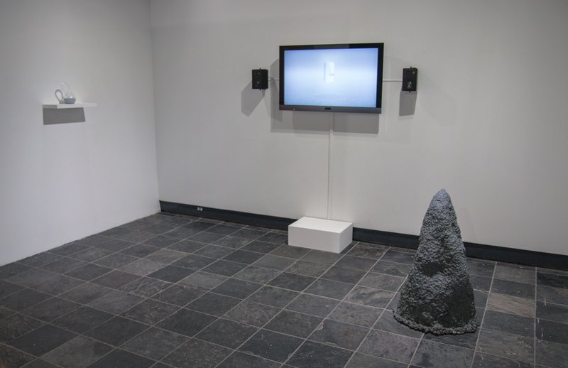 Art installation: TV screen with white image, black speakers, white shelf with glass, and dark gray conical sculpture on tiled floor.