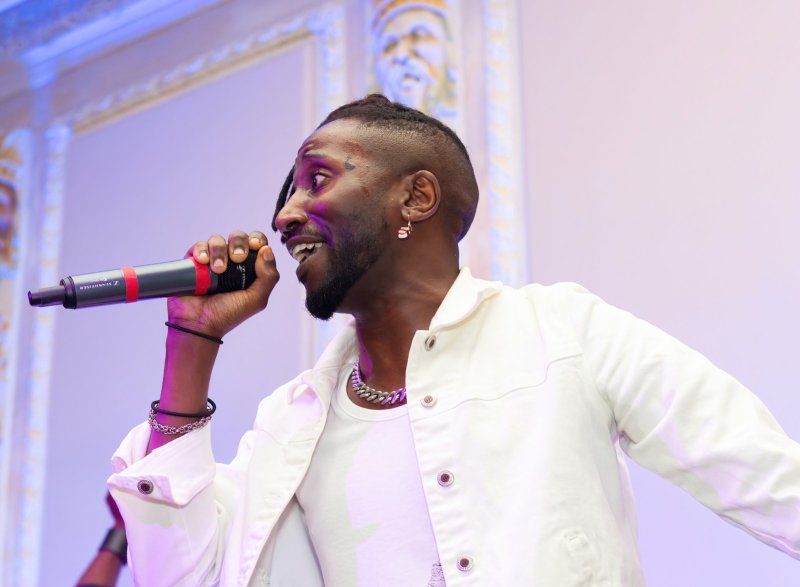 A person of color sings into a microphone, wearing a white outfit and silver chain. The background is a soft purple with architectural details visible.