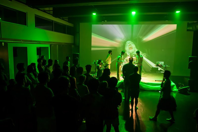 Audience watches four performers in green light before a large screen showing a distorted face screaming into a tunnel.