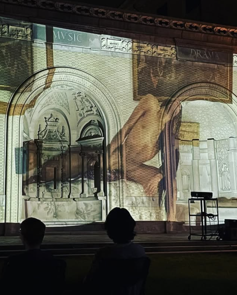 Projection of a large, nude figure onto a brick building with arched windows, viewed from behind by two silhouetted people.