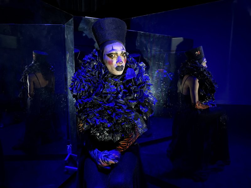 Drag performer in black and orange costume, clown makeup, and tall hat sits between mirrors reflecting their image in blue light.