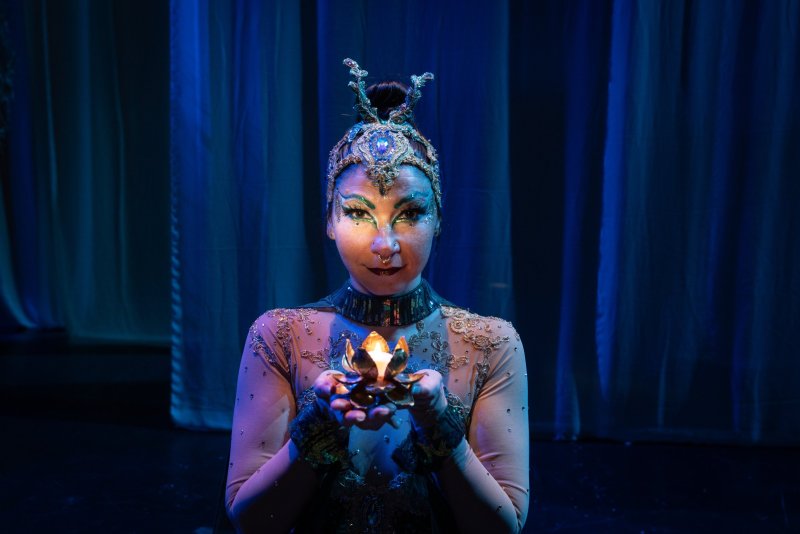 Woman in elaborate dragonfly-inspired costume and makeup, holding a glowing lotus, against a dark blue draped background.