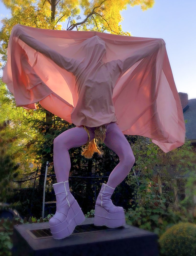 Figure in lavender tights and platform boots, draped in a pink cloth, against a vibrant autumn tree.