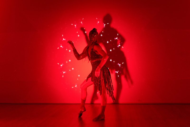 Dancer in a sparkling costume, striking a pose against a vibrant red background with confetti-like specks and a prominent shadow.