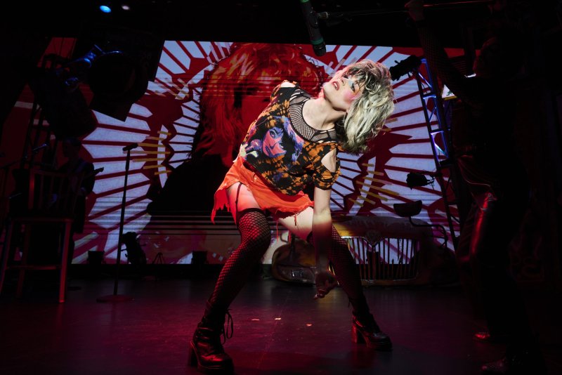 Performer in blonde wig, fishnets, ripped orange skirt, and black top, leaning back on a stage with a graphic red and white backdrop.